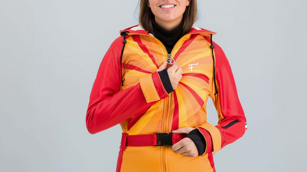 Femme souriante portant une combinaison de ski Hélios rouge-orange, inspirée des années 80, sur fond gris.