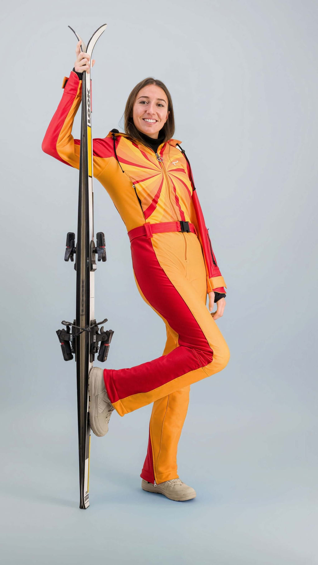 Femme portant une combinaison de ski Hélios rouge-orange avec des skis sur un fond neutre.